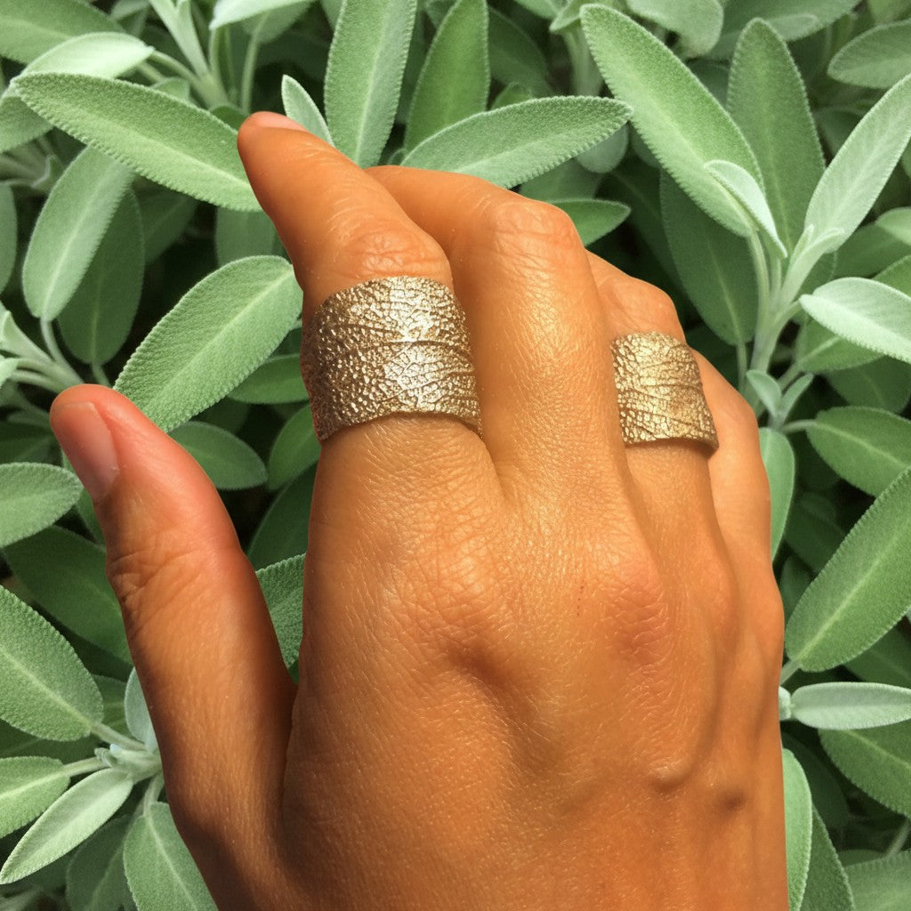 Anello MandalaT a forma di foglia di salvia con trama dettagliata indossato su una mano davanti a foglie di salvia