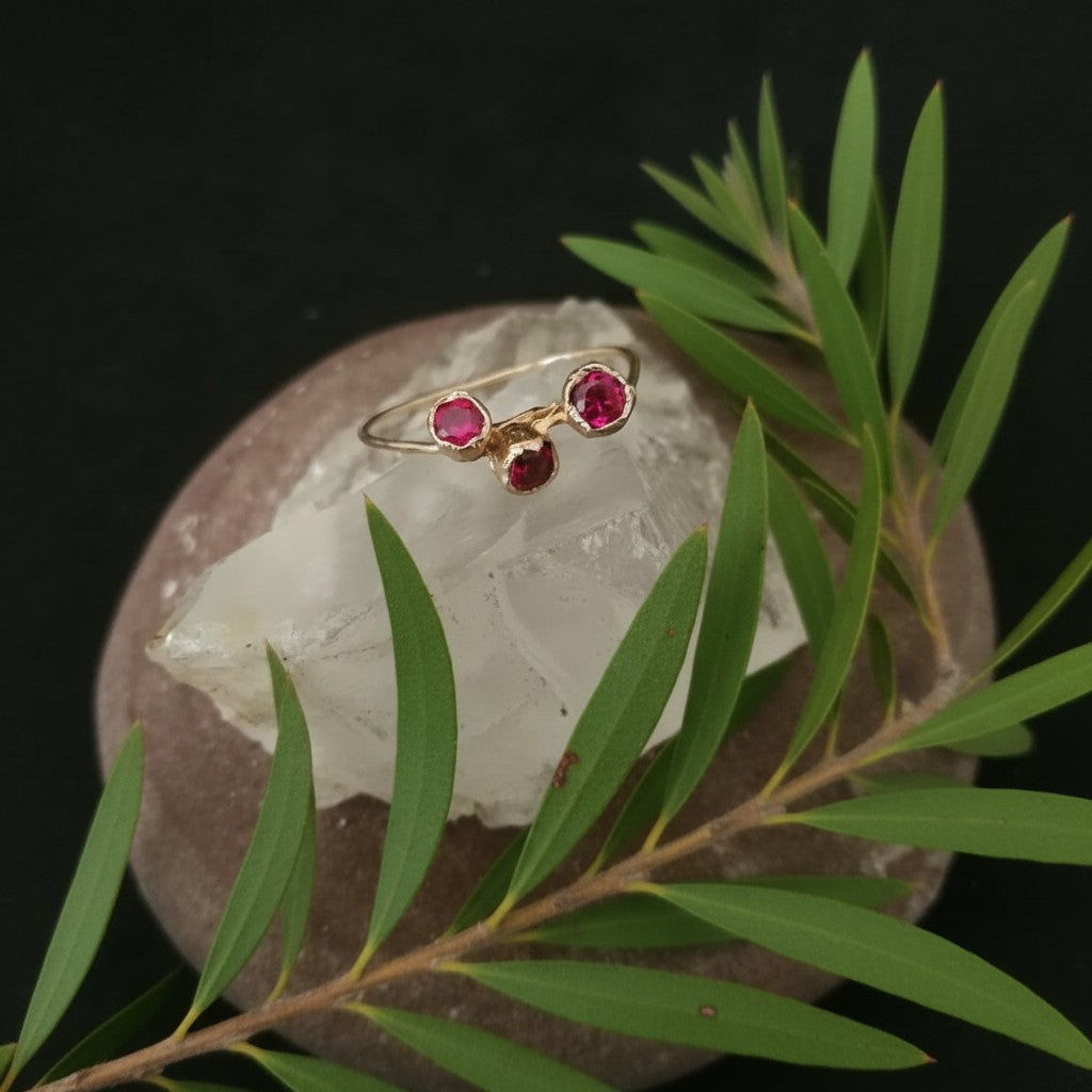 Anello Callistemone MandalaT in oro con tre rubini sintetici incastonate, poggiato su un cristallo e foglie verdi.