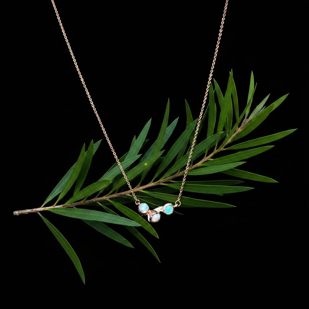 Collana MandalaT Callistemone in oro con tre pietre luminose ispirate ai fiori, su sfondo nero con ramo verde