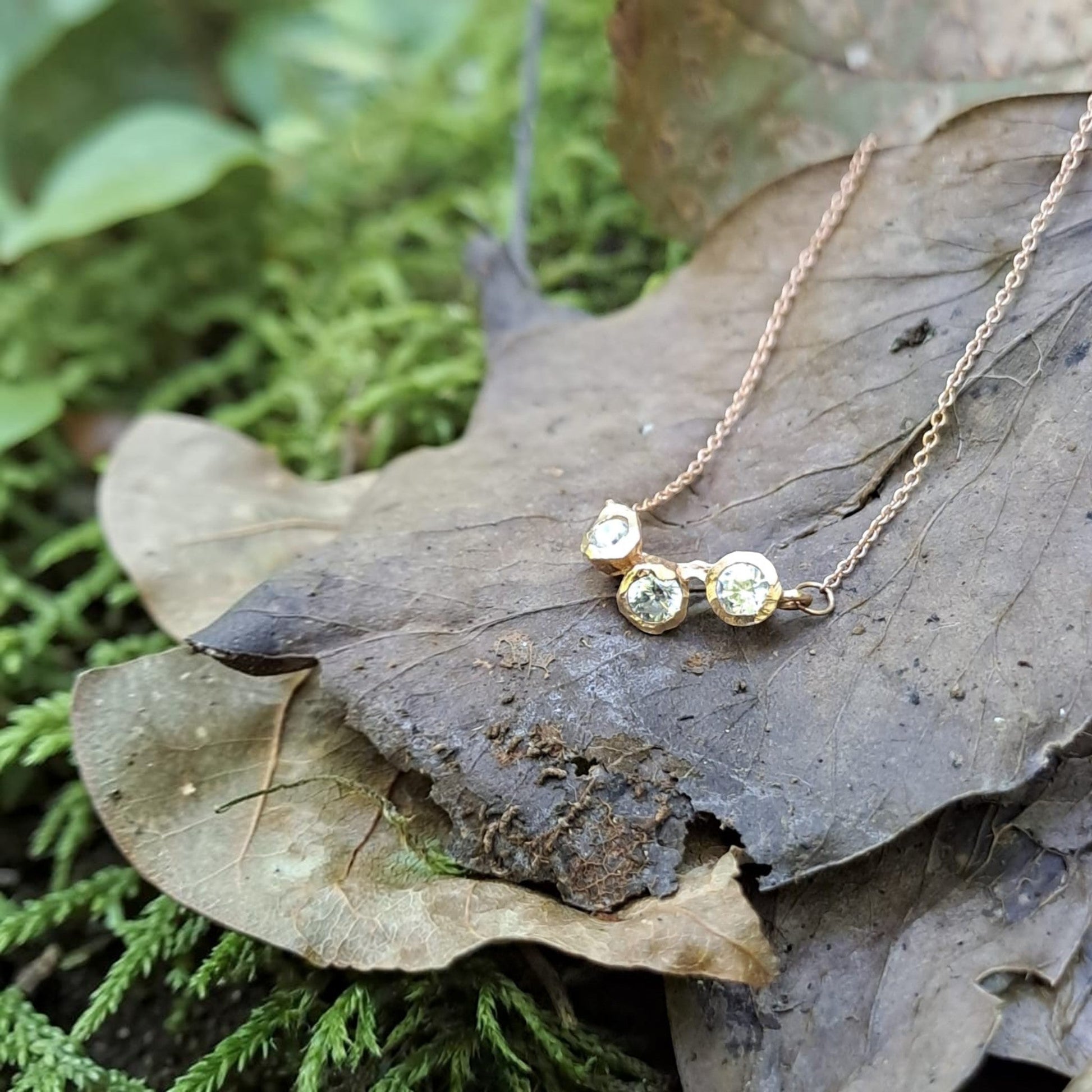 Collana MandalaT Callistemone in oro rosa con tre pietre luminose incastonate su una foglia secca nel bosco