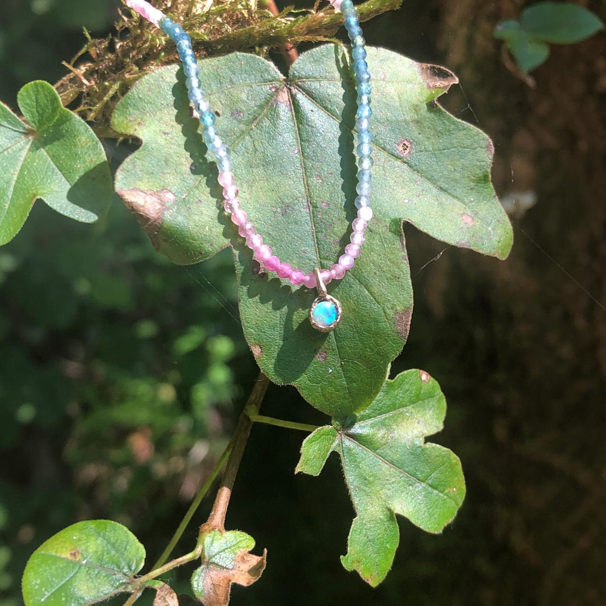 Collana MandalaT Callistemone con pietre sfaccettate rosa e azzurre e un ciondolo luminoso su una foglia verde