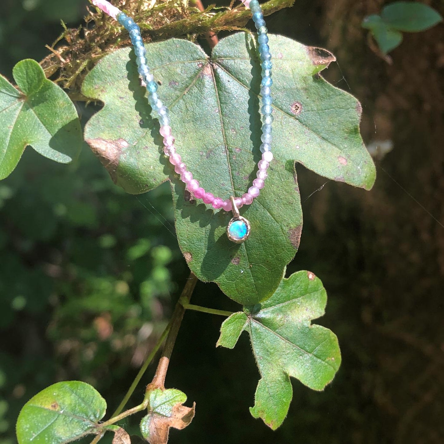 Collana MandalaT Callistemone con pietre sfaccettate rosa e azzurre e un ciondolo luminoso su una foglia verde