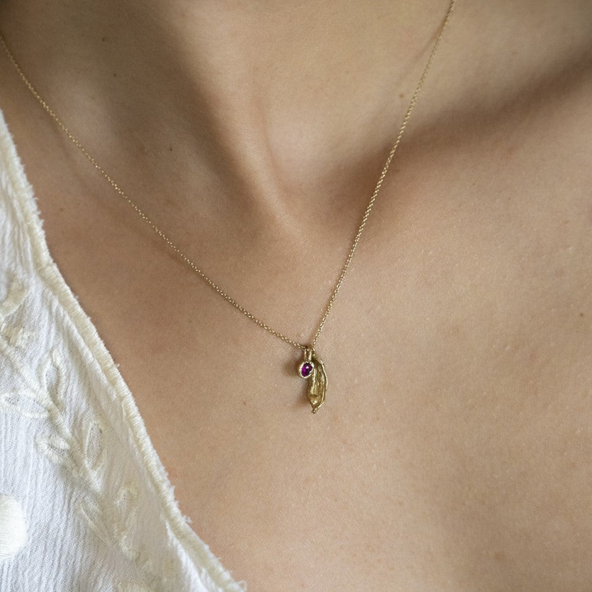 Collana MandalaT Cardamomo in oro con ciondolo a forma di baccello di Cardamomo e piccola pietra preziosa fucsia indossata dentro una fusione del fiore del Callistemone