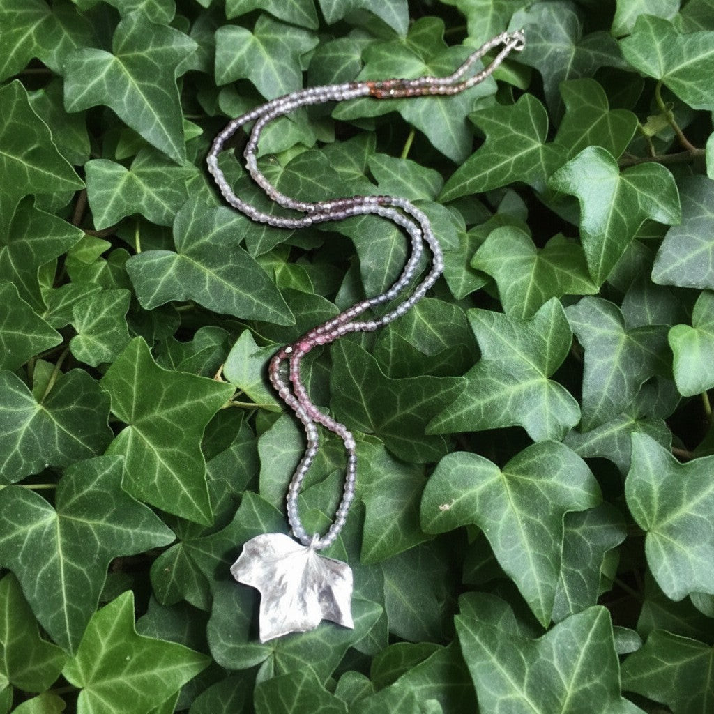 Collana Edera MandalaT con ciondolo a foglia in argento e catenina di perline sfumate su sfondo di foglie vere
