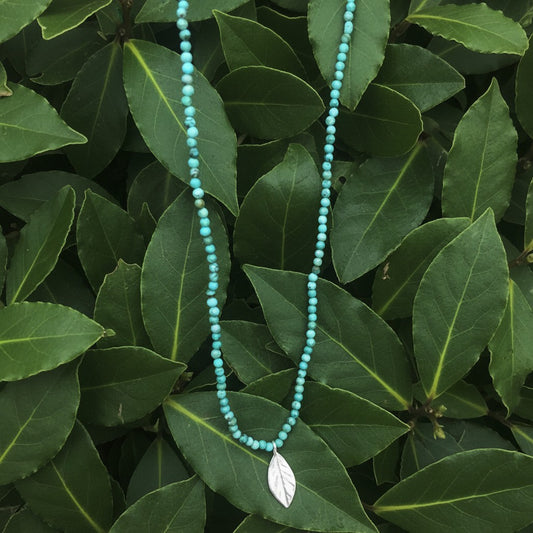 Collana MandalaT con perline turchesi e ciondolo a forma di foglia di alloro in argento su sfondo di foglie verdi