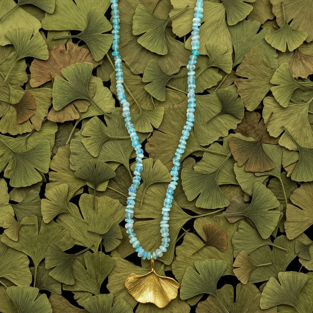 Collana MandalaT Gingko con ciondolo a foglia dorata e pietre azzurre su sfondo di foglie di ginkgo