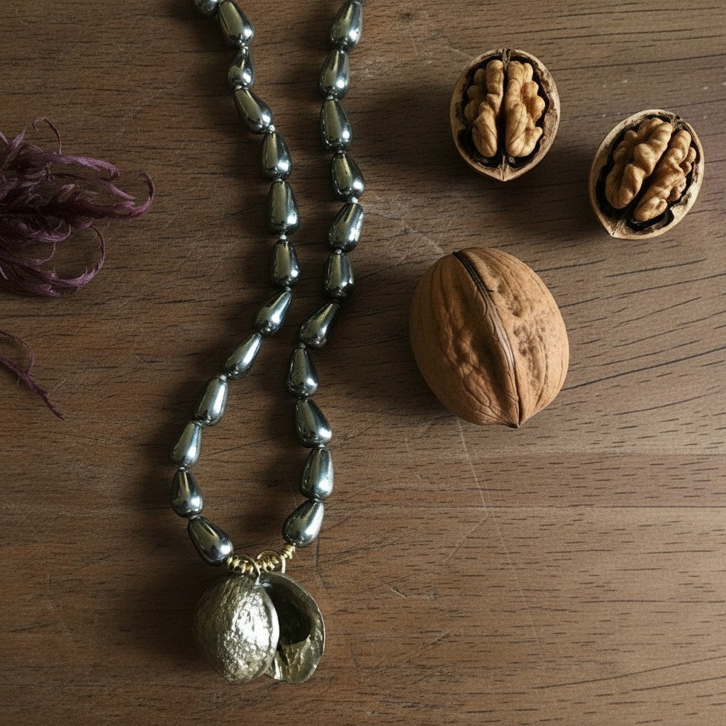 Collana MandalaT con pendente a forma di noce dorata e catena di perle metalliche grigie su sfondo in legno