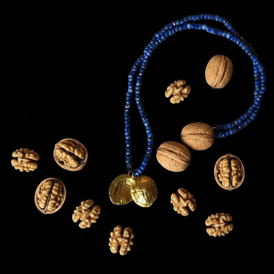 Collana MandalaT Noce con pendente dorato e catena di perline blu, circondata da noci su sfondo nero.