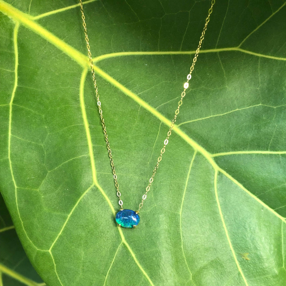 Collana MandalaT con ciondolo in opale blu ovale su sottile catenina d'oro, appoggiata su una foglia verde.