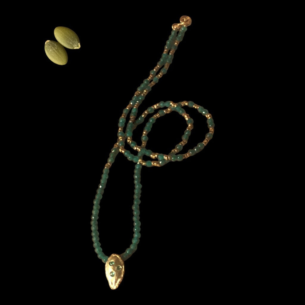 Collana MandalaT con ciondolo a forma di seme di zucca dorato e catenina di perline verdi e bronzo su sfondo nero