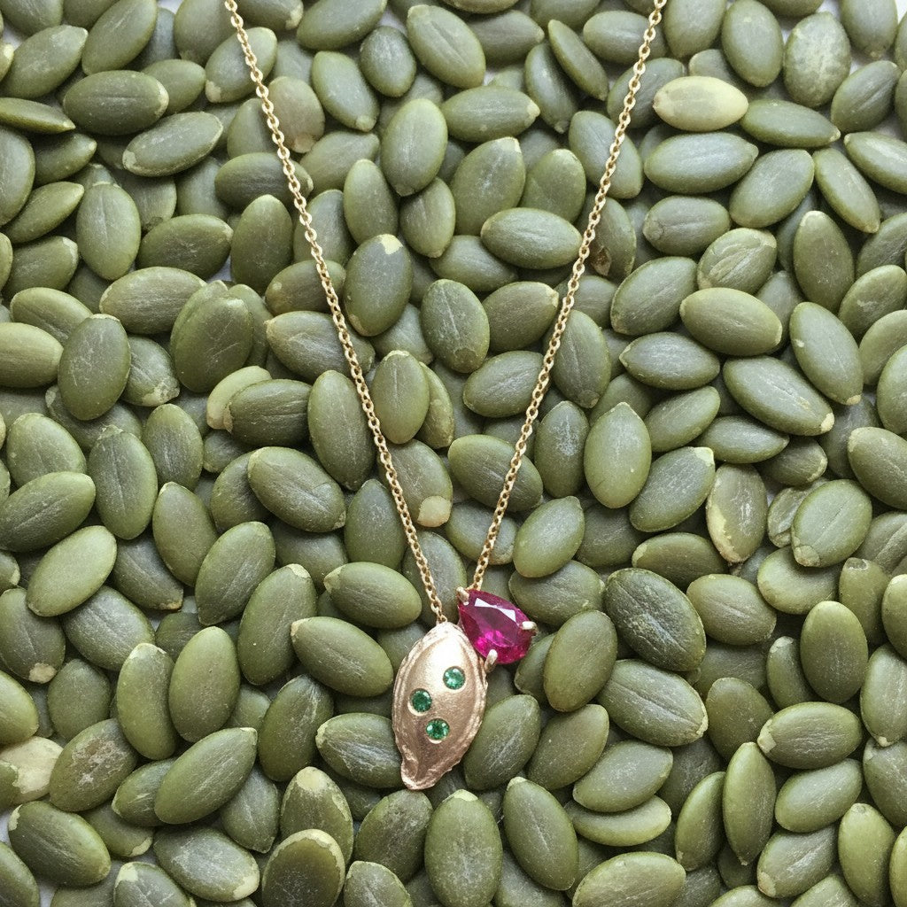 Collana MandalaT in oro con ciondolo a forma di seme di zucca, impreziosito da pietre preziose verdi e rosa.