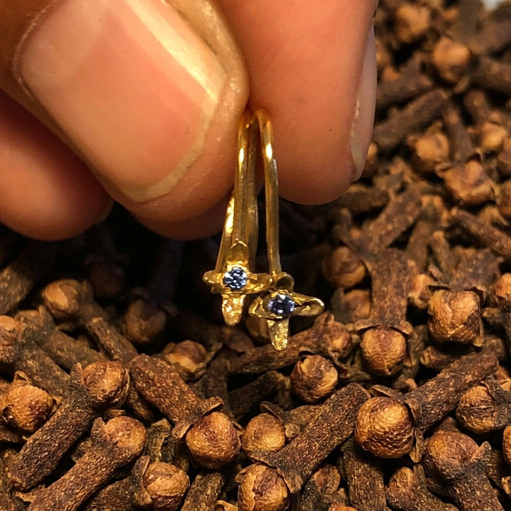 Orecchini ad amo MandalaT Chiodo di garofano in oro con piccola pietra preziosa blu, tenuti tra le dita.