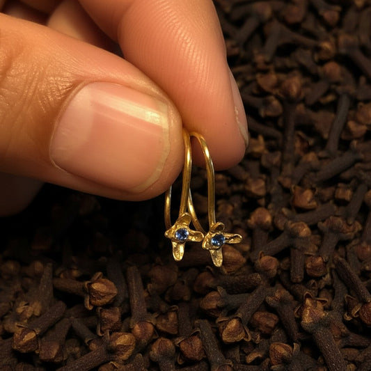 Orecchini ad amo MandalaT Chiodo di garofano in oro con pietra preziosa blu, tenuti tra le dita su sfondo di chiodi