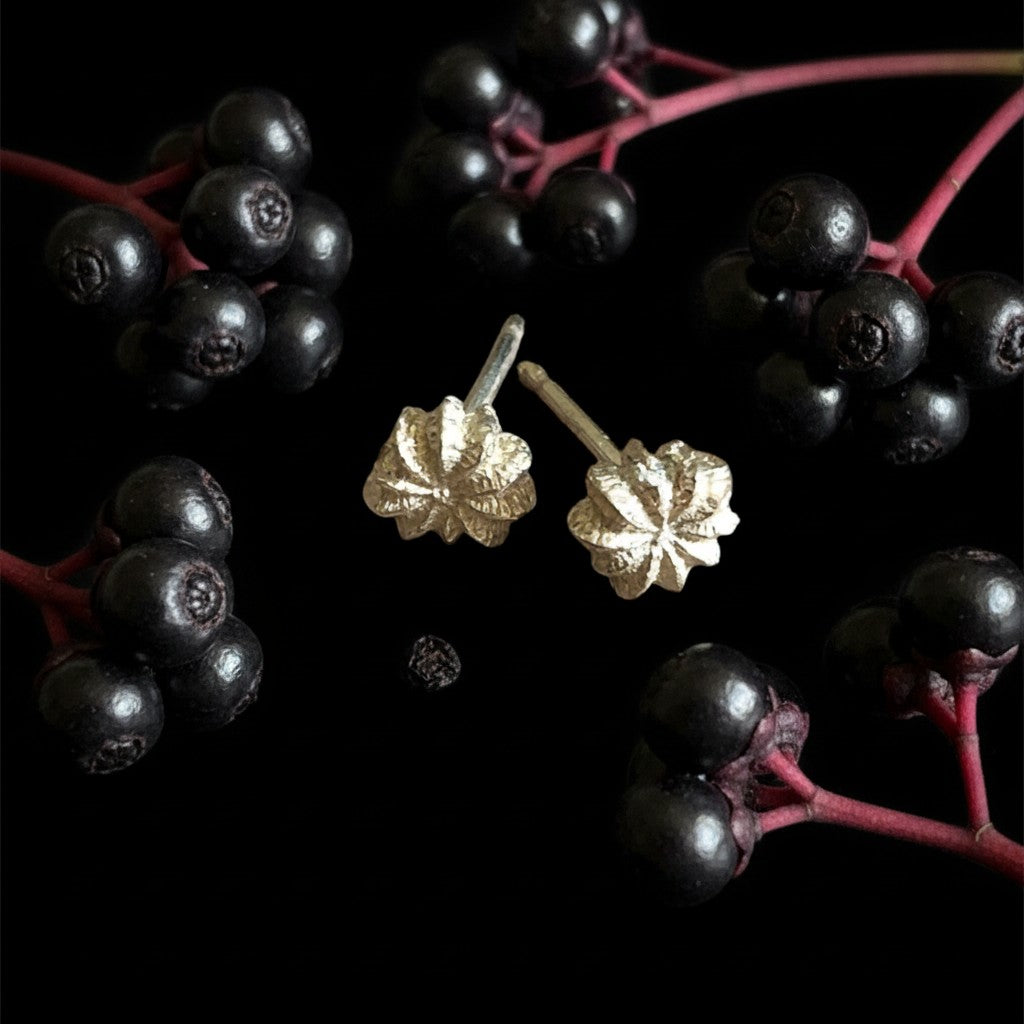 Orecchini a lobo MandalaT Phytolacca in argento a forma di fiorellino, posizionati tra bacche scure su sfondo nero.